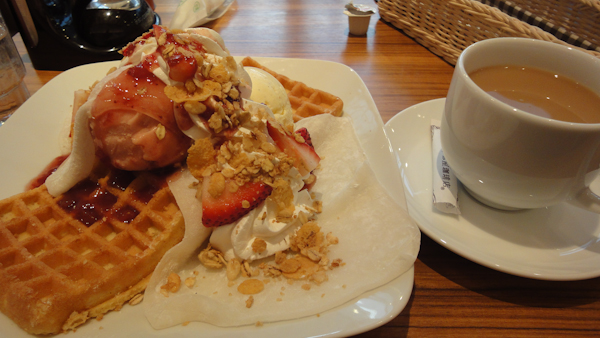 a cup of coffee and a plate of waffles topped with scoops of strawberry ice cream, sheets of mochi, whipped cream, and sliced almonds
